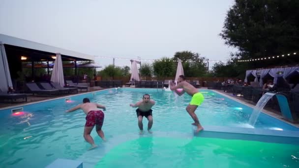 Gelukkige vrienden springen en spetteren in het zwembad in luxe resort. Jonge mannen in zwemkleding die 's nachts feesten in een privé vakantievilla. Jongens die in het water duiken. Langzame beweging. — Stockvideo