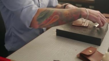 Deri ustası atölyesinde çalışıyor. Usta deri cüzdan yapıyor. Adam delen deri yumruk aletleriyle delik açıyor. Yumruk ve çekiç dikiyor..