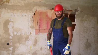 Müteahhit ayakta duruyor, parçalamak ve yıkmak için balyozu omuza kaldırıyor. Koruyucu gözlüklü, koruyucu şapkalı, tulumlu, kameraya bakan kendine güvenen bir inşaat işçisi. Yavaş çekim.
