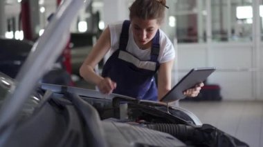 Kadın tamirci kaputun altındaki araba parçalarını inceliyor. Tulumlu kadın motoru kontrol ediyor ve elinde kontrol listesi ve talimatlarla dijital tablet tutuyor. Modern bir atölyede araç tamiri hatası.