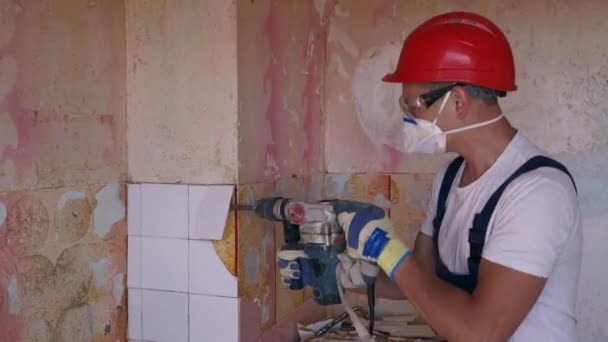 Contratista elimina de baldosas cerámicas frente a la pared. Trabajador haciendo trabajos de desmontaje y demolición con taladro de martillo rotatorio. Trabajos de renovación de apartamentos. Constructor en uniforme y de trabajo hardhat. — Vídeos de Stock