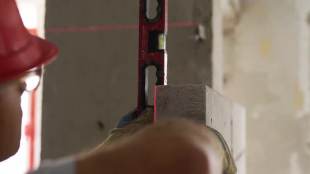 Murare med hjälp av vattenbubbla och lasernivå för att exakt kontrollera betongblock på väggen. Entreprenören använder verktyg för tegel. Arbetstagare bygger en vägg i ny lägenhet fastigheter. — Stockvideo