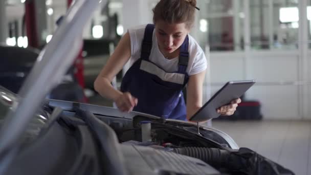 Ženský mechanik kontrolující komponenty pod kapotou. Žena v montérkách zkontroluje motor a drží digitální tablet s kontrolním seznamem a instrukcemi. Upevnění poruchy vozidla v moderní dílně. — Stock video