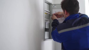 Elektrikçi elektrik sigorta kutusunu söker. Üniformalı usta elektrik panelinin vidalarını tornavidayla söküp elektriği tamir ediyor. Tamirci ev içi sorunları düzeltiyor.