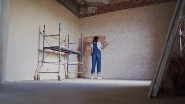 Profesyonel alçı dişisi bir duvarı alçılıyor, alçıyı tulum ve beyzbol şapkasıyla bir spatulaya sürüyor ve yayıyor. Duvarları sıva kaplı genç bir kadın, inşaat alanında dekorasyon işi yapıyor. İşlemler.