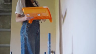 Profesyonel ressam kadın, inşaat alanında duvarı silindir ve tepsiyle boyuyor. Tulumlu ve şapkalı genç bir kadın ev dekorasyonu ve tadilat yapıyor. İşçi bir odada DIY onarımı yaptırıyor.