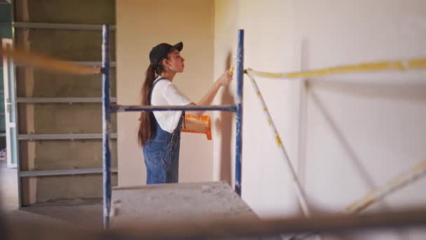 La pintora profesional pinta la pared con rodillo y bandeja en el sitio de construcción. Mujer joven en overoles y gorra haciendo decoración y renovación del hogar. Trabajador hace trabajos de reparación de bricolaje en la habitación. — Vídeos de Stock