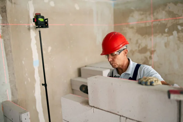 Bricklayer using rubber mallet hammer to tap, level concrete blocks wall. Contractor uses precise spirit bubble and laser level for brickwork. Worker constructs a wall in new apartment real estate.