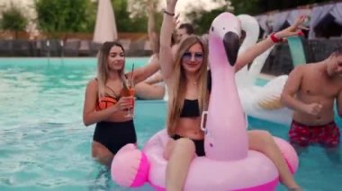 Gece havuzu partisinde şişme pembe flamingo şiltede takılan bikinili güzel bir kadın. Arkadaşlar tatil köşkünde kokteyllerle parti yapıyorlar..