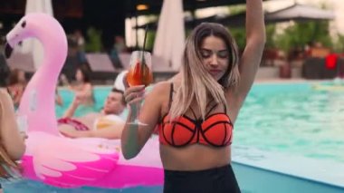 Bikinili güzel bir kadın kokteyl içiyor ve gece havuzu partisinde arkadaşlarıyla takılıyor. İnsanlar lüks bir yüzme havuzunda içkilerle dans edip su sıçratarak eğleniyorlar..