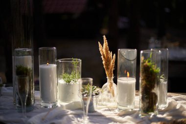 Düğün partisi dışarıda ormanda ziyafet veriyor. Mum, beyaz kumaş, çiçek, tabak, sofra takımı, yemek takımı, yemek takımı, kadeh ve içeceklerle süslenmiş yemek masası. Tatil yemekleri.