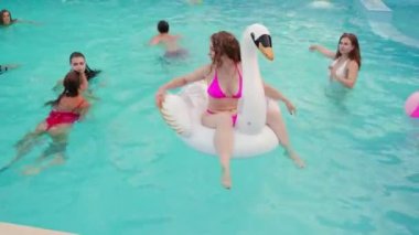 Gece havuzu partisinde bikinili güzel bir kadın şişme beyaz kuğu yatağında takılıyor. Arkadaşlar tatil köşkünde kokteyllerle parti yapıyorlar..