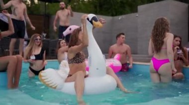 Gece havuzu partisinde şişme beyaz kuğu şiltesine takılan gerçek bikinili yaşlı bir kadın. Arkadaşlar tatil köşkünde kokteyllerle parti yapıyorlar..