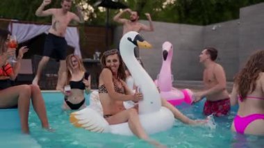 Gece havuzu partisinde şişme beyaz kuğu şiltesine takılan gerçek bikinili yaşlı bir kadın. Arkadaşlar tatil köşkünde kokteyllerle parti yapıyorlar..