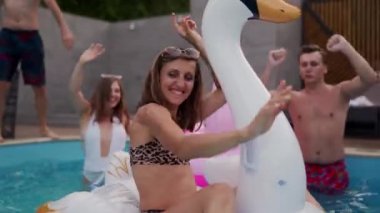 Gece havuzu partisinde şişme beyaz kuğu şiltesine takılan gerçek bikinili yaşlı bir kadın. Arkadaşlar tatil köşkünde kokteyllerle parti yapıyorlar..