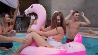 Gece havuzu partisinde şişme pembe flamingo şiltede takılan bikinili güzel bir kadın. Arkadaşlar tatil köşkünde kokteyllerle parti yapıyorlar..