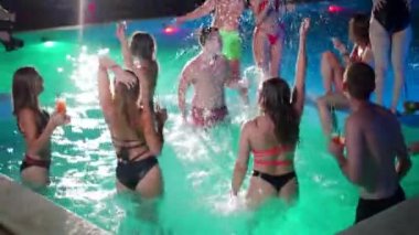 Gece havuzu partisinde yüzme havuzunda su sıçratan arkadaşlar. Mayo giyen, dans eden, lüks bir tatil köyünde parti yapan neşeli gençler. Mutlu erkekler ve kadınlar birlikte takılıyor. Yavaş çekim.