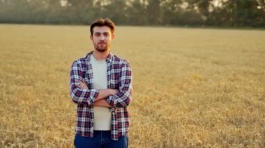 Olgun buğday tarlasında kolları göğsünde duran mutlu çiftçinin portresi. Çiftlik arazisindeki kameraya bakan gururlu tarım uzmanı. Zengin ekili mısır gevreği melezi hasadı. Hasat mevsimi.