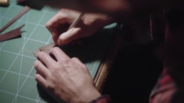 Yakın plan eller bıçakla deri desenleri oyar ve fazlalık kısımları çıkartır. Tanner, terzi tezgahın arkasında çalışıyor ve deri eşyalar yapıyor..