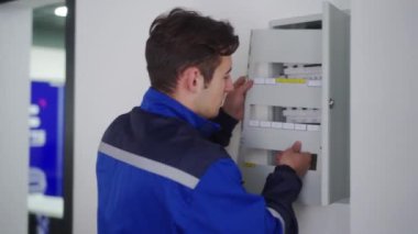 Elektrikçi bir elektrik sigorta kutusunu söküyor. Üniformalı usta elektriği tamir etmek için elektrik panelini erteliyor. Tamirci şalterleri ve şalterleri denetleyerek iç sorunları düzeltiyor..