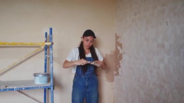 Profesyonel alçı dişisi bir duvarı alçılıyor, alçıyı tulum ve beyzbol şapkasıyla bir spatulaya sürüyor ve yayıyor. Duvarları sıva kaplı genç bir kadın, inşaat alanında dekorasyon işi yapıyor. İşlemler.