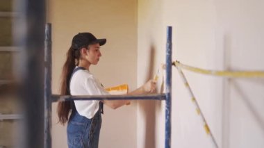 Profesyonel ressam kadın, inşaat alanında duvarı silindir ve tepsiyle boyuyor. Tulumlu ve şapkalı genç bir kadın ev dekorasyonu ve tadilat yapıyor. İşçi bir odada DIY onarımı yaptırıyor.