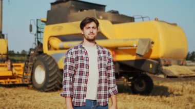 Göğsünde elleri çapraz olan gururlu hasatçı makinist portresi. Çiftçi kombinasyonunda duruyor. Agronomist kameraya bakıyor. Hasat edilen buğday tarlaları üzerinde çalışan çiftçi..