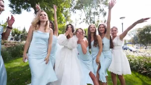 漂亮的新娘和漂亮的伴娘们穿着浅蓝色的衣服 在公园里挥手欢呼 穿着白色婚纱 头戴长袖婚纱的女人 请与女友们亲热 — 图库视频影像