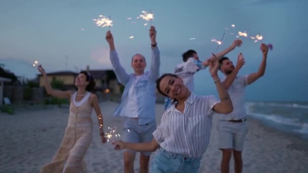 7月4日には 海沿いのナイトパーティーを楽しみながら 多民族の友人たちが歩き 輝く光を手にしています 新年やクリスマスに花火でビーチでパーティーをする若者たち — ストック動画