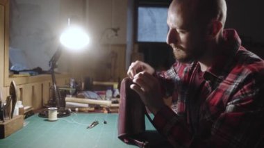 Deri ustası el çantası yapıyor. İğne iplikli adam deri atölyesinde çalışıyor. Ekose gömlekli terzi çanta yapıyor otantik atölyeye aksesuar dikiyor..