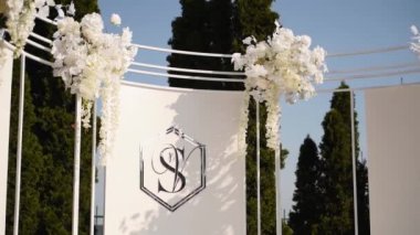 Nişanlı düğün kemeri arka planında gelin ve damat isminin baş harfleri var. Beyaz çiçeklerle süslenmiş düğün kemeri, açık havada yeşillik. Yeni evlilerin aile logosu. İsimlerin ilk harfleri..
