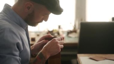 Deri ustası bir atölyede çalışıyor. Usta deri cüzdan yapıyor. İğne iplikli bir adam atölyedeki tezgahta dikiş malzemeleri ve aksesuarlar dikiyor..