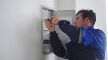 Elektrikçi bir elektrik sigorta kutusunu söküyor. Üniformalı usta elektrik panelinin vidalarını tornavidayla söküp elektriği tamir ediyor. Tamirci ev içi sorunları düzeltiyor.
