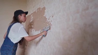 Profesyonel alçı dişisi bir duvarı alçılıyor, alçıyı tulum ve beyzbol şapkasıyla bir spatulaya sürüyor ve yayıyor. Duvarları sıva kaplı genç bir kadın, inşaat alanında dekorasyon işi yapıyor. İşlemler.
