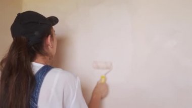 Profesyonel ressam kadın, inşaat alanında duvarı silindir ve tepsiyle boyuyor. Tulumlu ve şapkalı genç bir kadın ev dekorasyonu ve tadilat yapıyor. İşçi bir odada DIY onarımı yaptırıyor.