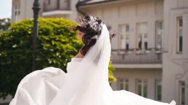 Uzun, havadar peçeli güzel bir gelin, lüks bir saray bahçesinde düğün günü havada uçuşan kumaşla dönen zarif beyaz bir elbise. Çekici, çekici bir kadın kameraya poz veriyor, muhteşem bir elbise içinde..