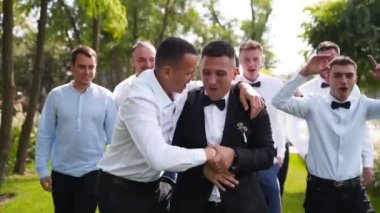 Damat esnetiyor ve düğün günü sağdıçlarla yürüyüş yapıyor. Siyah takım elbiseli, papyonlu ve yaka çiçekli mutlu bir adam arkadaşlarıyla takılıyor ve parkta kameraya poz veriyor..