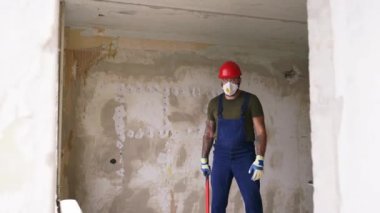 Müteahhit ayakta duruyor, yıkımı yeniden düzenlemeden önce dairede balyoz kaldırıyor. Koruyucu gözlüklü, şapkalı, tulumlu, kameraya bakan inşaat işçisi. Kahraman vuruşu..