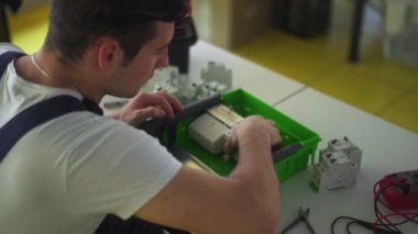 Elektrikçi, elektrik paneli sigorta kutusunu atölyede monte ediyor. İşçi masadaki dağıtım panosuna enerji sayacı, şalterler ve devre kesiciler yerleştirir.