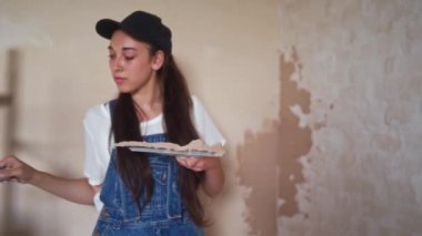 Profesyonel alçı dişisi bir duvarı alçılıyor, alçıyı tulum ve beyzbol şapkasıyla bir spatulaya sürüyor ve yayıyor. Duvarları sıva kaplı genç bir kadın, inşaat alanında dekorasyon işi yapıyor. İşlemler.