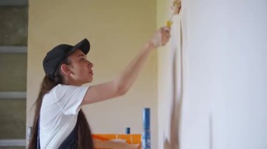 Profesyonel ressam kadın, inşaat alanında duvarı silindir ve tepsiyle boyuyor. Tulumlu ve şapkalı genç bir kadın ev dekorasyonu ve tadilat yapıyor. İşçi bir odada DIY onarımı yaptırıyor.