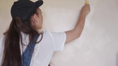 Profesyonel ressam kadın, inşaat alanında duvarı silindir ve tepsiyle boyuyor. Tulumlu ve şapkalı genç bir kadın ev dekorasyonu ve tadilat yapıyor. İşçi bir odada DIY onarımı yaptırıyor.