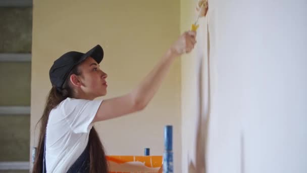专业画家女性在建筑工地上用滚子和托盘在墙上作画 穿着工作服 头戴帽子的年轻女子在做家居装饰和装修 工人们在一个房间里做Diy修理工作 — 图库视频影像