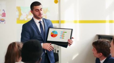 Businessman presents report, shows diagrams on tablet pc in conference room for multiethnic partners. Diverse international team listen CEO on board meeting in office. Corporate people discuss a plan.