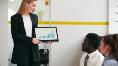 Business woman presents report, shows diagrams on tablet pc in conference room for multiethnic partners. Diverse international team listen CEO on board meeting in office. Corporate people discuss a