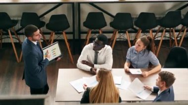Businessman presents report, shows diagrams on tablet pc in conference room for multiethnic partners. Diverse international team listen CEO on board meeting in office. Corporate people discuss a plan.