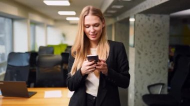 Smiling business woman using mobile phone, typing text messages standing in an office. Portrait of female texting with smartphone application. Blonde girl in smart casual wear. Internet communication.
