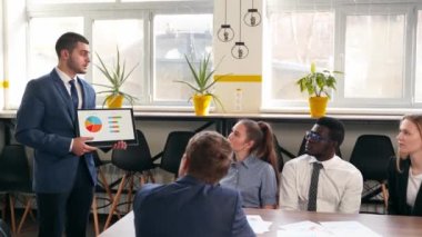 Businessman presents report, shows diagrams on tablet pc in conference room for multiethnic partners. Diverse international team listen CEO on board meeting in office. Corporate people discuss a plan.