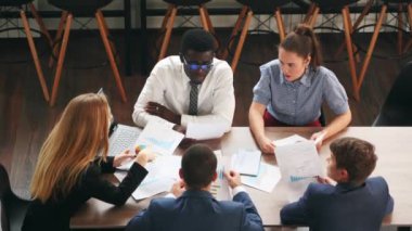 International business partners discuss financial project results on meeting in office. Multiethnic corporate team plans marketing strategy with diagrams, charts, papers, devices. People brainstorming