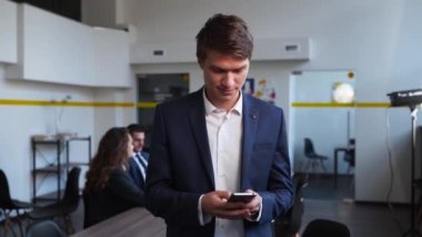 Smiling eastern caucasian businessman using mobile phone, typing text messages standing in an office. Portrait of male texting with smartphone application. Guy in formal wear communicating with
